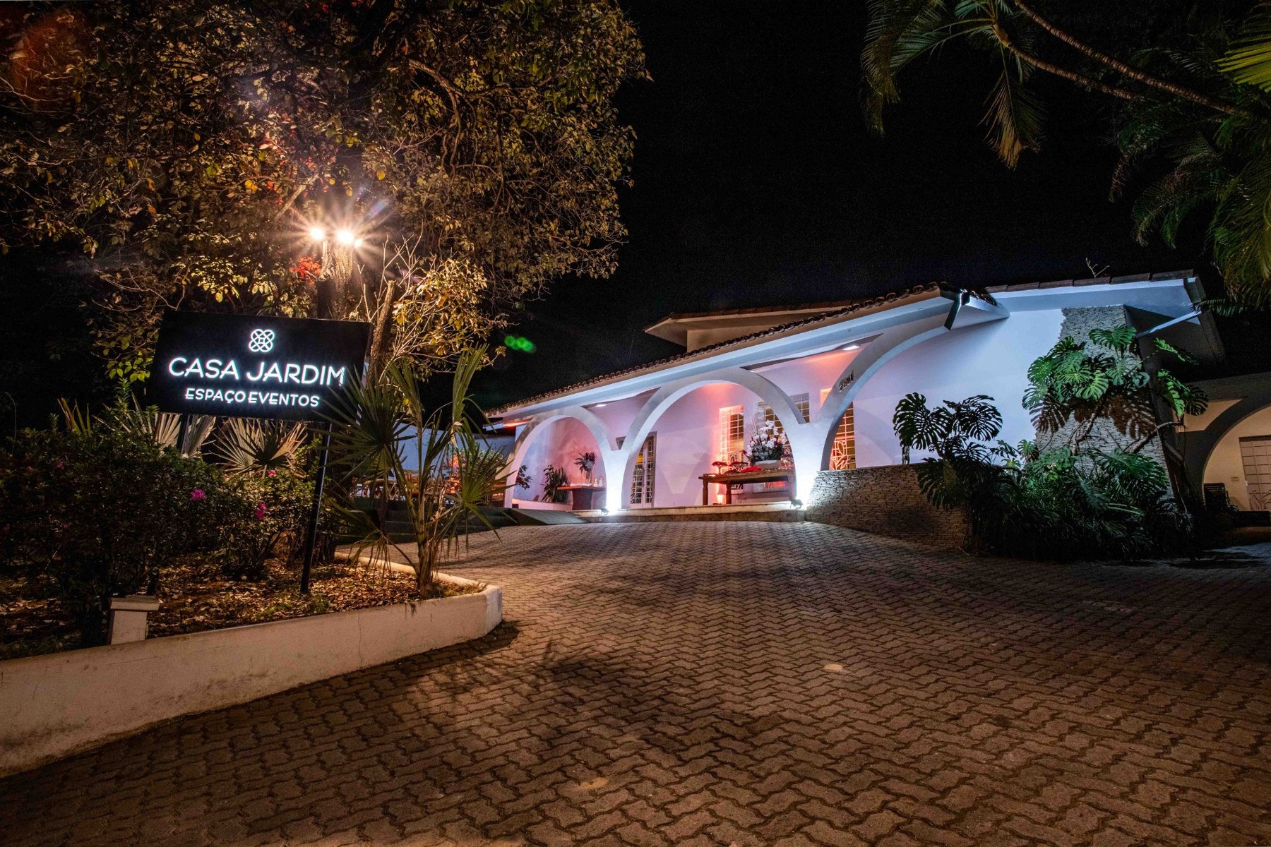 Fachada iluminada da Casa Jardim, entre árvores e jardins